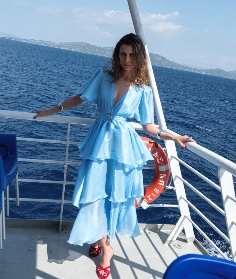 Ολόσωμη μπροστινή όψη με το μοντέλο να φοράει το γαλάζιο φόρεμα Nazezhda Sarah Dress Baby Blue από μεταλλική οργάντζα, αναδεικνύοντας το τριπλό βολάν.