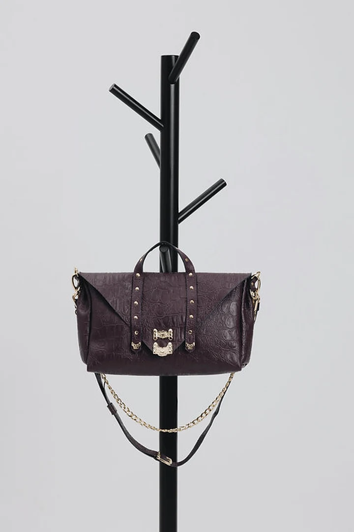 Studio φωτογραφία της τσάντας Individual Art Leather Surrender Dark Plum σε κρεμάστρα με τη χρυσή αλυσίδα και τον δερμάτινο ιμάντα