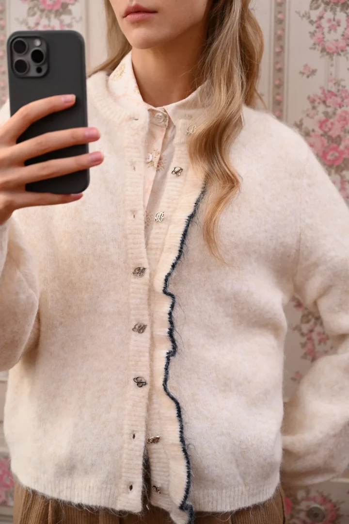 Κοντινό πλάνο στην εκρού χνουδωτή ζακέτα Boom Birdie Cardigan, αναδεικνύοντας τα μεταλλικά κουμπιά σε σχήμα B, τη χνουδωτή υφή και την κυματιστή πατιλέτα με σκούρο περίγραμμα