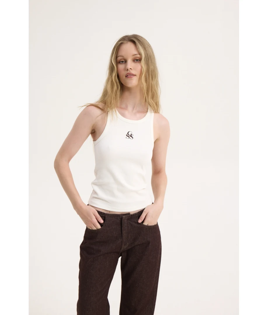Λήψη από τα 3/4 με το μοντέλο να φοράει το off-white ριμπ αμάνικο τοπ SSG Tank Top της Sunsetgo, με τα χέρια στις τσέπες του παντελονιού
